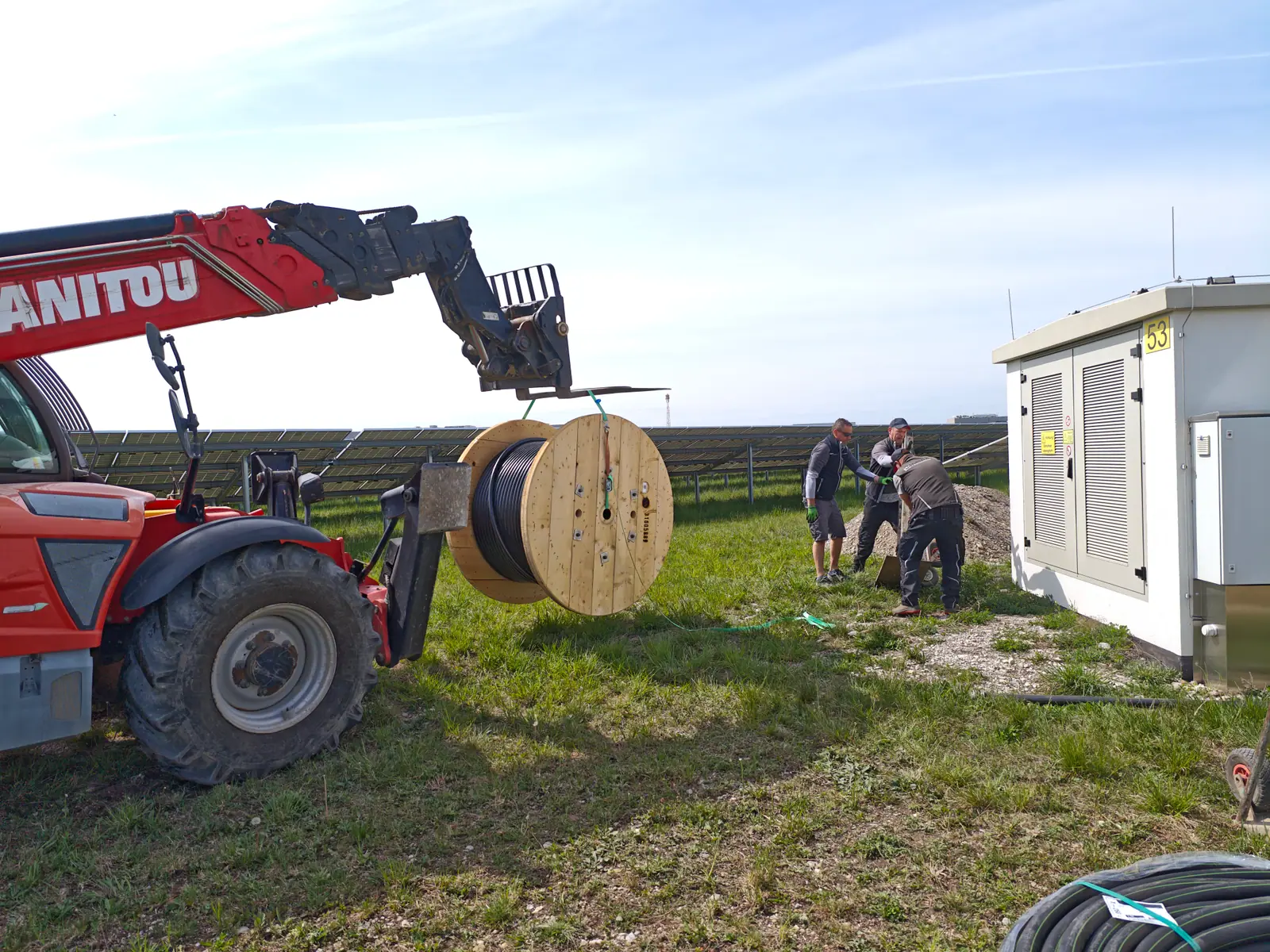Errichtung im Solarpark — Manitou-Lader mit Kabeltrommel und Techniker beim Trafohaus zwischen den PV-Reihen